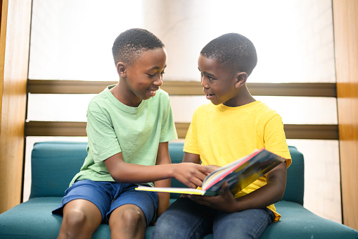 Two young boys sitting in the library looking & talking about the book they are reading