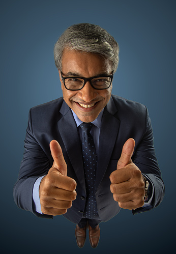 High angle portrait of confident positive senior businessman showing thumbs up gesture while standing against blue background