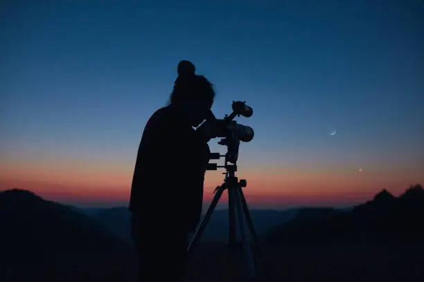 Photo of Astronomer looking at the starry skies and crescent Moon with a telescope.