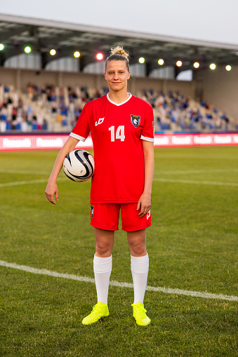 Portrait of smiling female football player in red jersey standing on football pitch during match.