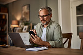 A smiling male freelancer with eyeglasses using a phone while working over a laptop.