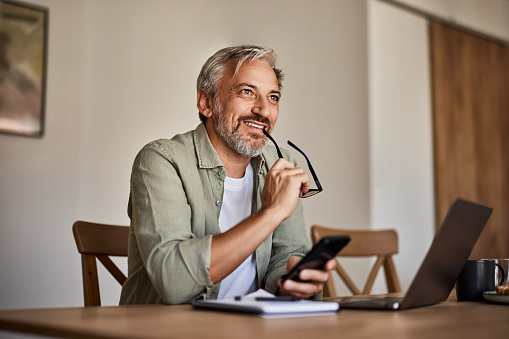 Handsome mature man looking at a distance holding a smartphone and eyeglasses, while sitting at wooden table with laptop and notebook.