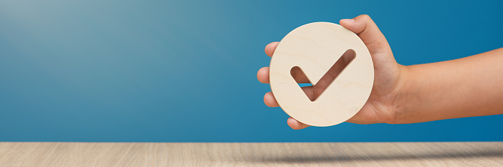 Hand holding check mark icon, banner with wooden check mark icon in hand, right sign. On a blue background. Copy space. Place for text. High quality photo