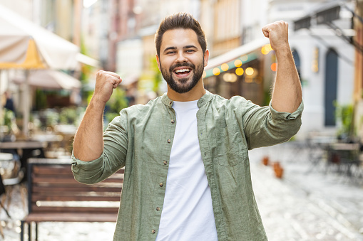 Bearded young man shouting, celebrating success, winning, goal achievement, good news, lottery jackpot luck, victory outdoors. Caucasian guy walking in urban city sunshine street. Town lifestyles