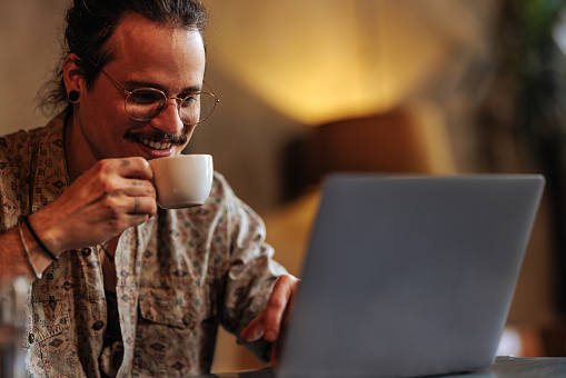 Hispanic adult man holding a cup of coffee while using a laptop in a cafe.
