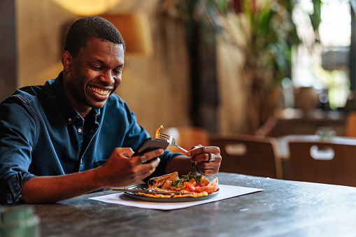 Young and happy African-American man is using a phone while having a lunch at the restaurant.