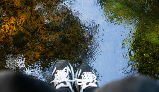 Women's legs hang over the river. Reflection of foliage and grass in the water.