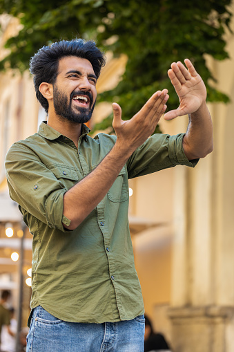 Cheerful rich bearded indian man showing wasting throwing money around hand gesture, more tips earnings, big profit, win lottery, share, celebrate outdoors. Guy standing in urban city street. Vertical