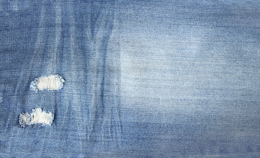 Ripped jeans material of indigo color. Horizontal or vertical background with denim texture of blue color with torn holes
