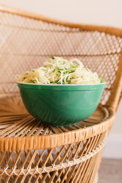 vibrant vintage teal borosilicate glass bowl filled with zucchini cut into spaghetti-shape sitting on a vintage rattan chair, ready for a healthy summertime dinner - crystal noodles imagens e fotografias de stock