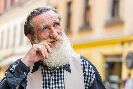 Portrait of happy bearded senior old man smiling friendly, fixing mustache, glad expression looking at camera, resting, relaxation feel satisfied good news outdoors. Mature grandfather in city street