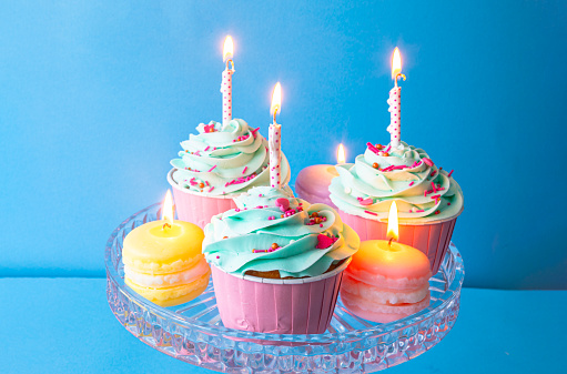 Birthday cupcakes on glass cake stand, with macaron candles, blue frosting, pink cases, and blue background