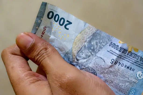A hand figure holding a two-thousand rupiah Indonesian banknote.