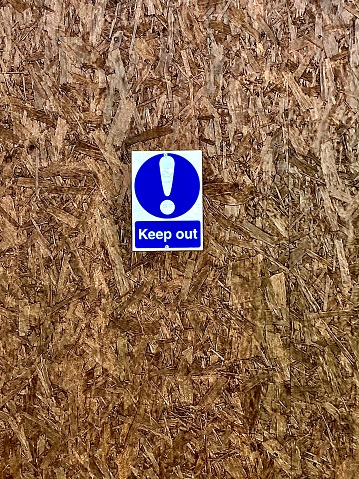 A small sign in the middle of medium brown chipboard, in blue and white, an exclamation mark with the words ‘keep out’ warning people not to trespass.