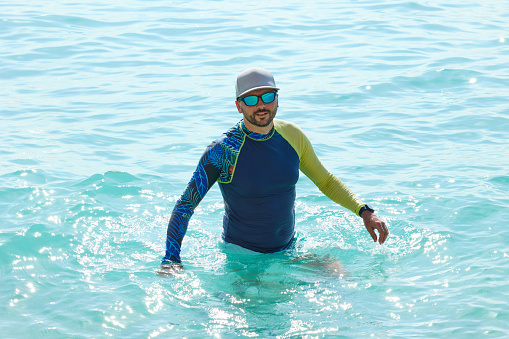 Handsome man with beard and mustache in a lycra shirt standing waist-deep in blue sea water.