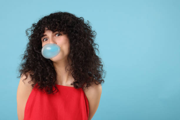 istock Beautiful young woman blowing bubble gum on light blue background. Space for text 1634418154