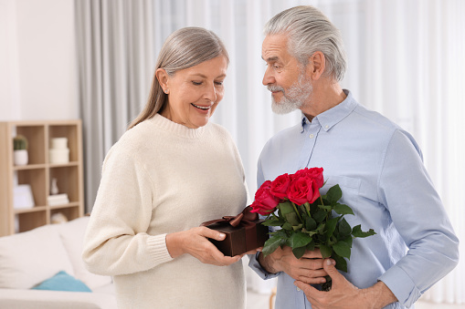 Happy affectionate senior couple with gift box and beautiful bouquet indoors