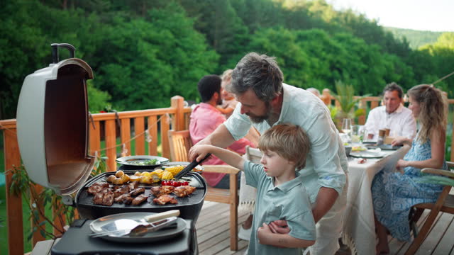 Father with little son grilling ribs and vegetable on grill during family summer garden party.