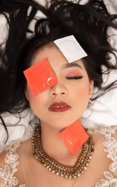 an Asian woman with a gold necklace falls asleep with a condom wrapper on her face in a hotel at night