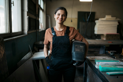 Portrait of a young woman welder at metal workshop. Happy female holding a welding protective helmet smiling at camera in metal workshop.