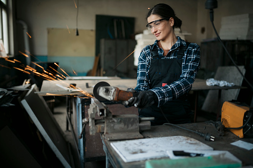 Young female apprentice wearing safety equipment operating a angle grinder on a workbench with metal held in a vice. Caucasian woman grinding a metal piece using angle grinder at small workshop.
