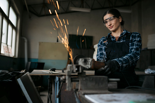 Female blacksmith cutting metal with angle grinder in workshop. Young woman worker using grinding machine to cut a metal piece over workbench.