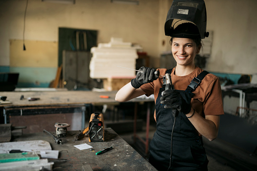 Portrait of a happy young woman welder with safety helmet with new electrode on arc welding holder in the workshop. Smiling female worker working with arc welding machine in workshop.