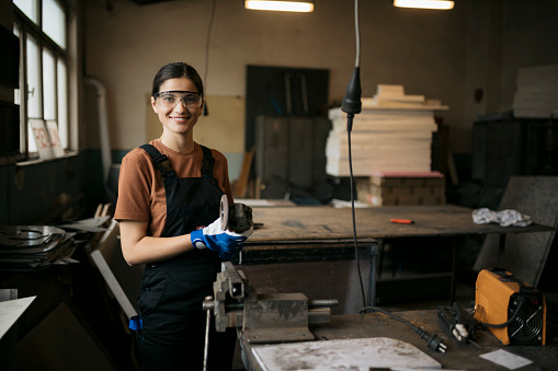 Portrait of young caucasian woman apprentice with safety equipment standing at workbench with a grinder in workshop. Young worker working in a small metal workshop looking at camera and smiling.