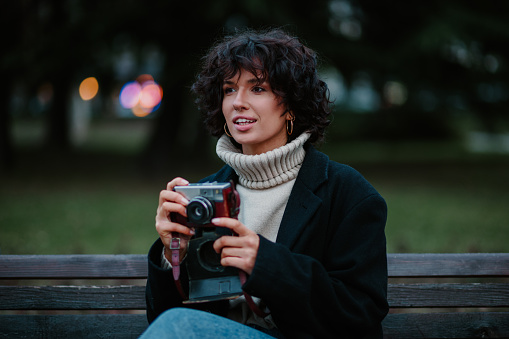 Cropped shot of a curious dark haired young woman sitting in a park on a bench and taking photos with an old retro camera. Looking into the distance while enjoying a beautiful winter day. Copy space.