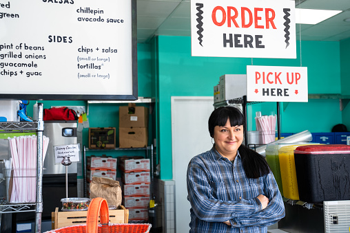 A mature business owner stands under the 'Order Here' sign in her restaurant.