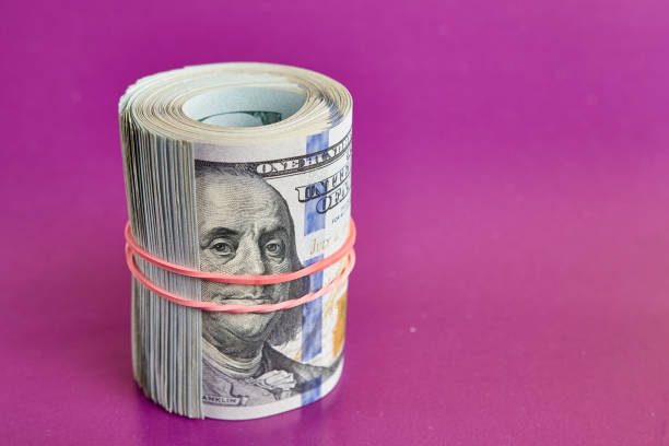 istock Many hundred dollar bills rolled up with a red rubber band on a pink background. 1635192258