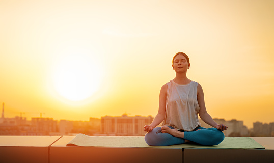 Young sportswoman is meditating on rooftop of parking, garage in summer day morning.