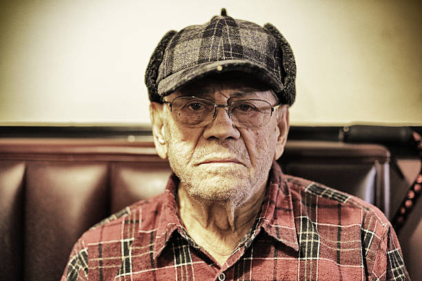 Portrait of a frowning 90 year old man sitting in a restaurant. He is dressed in a plaid hunters cap (with a tiny light on the bill) and a plaid lumberjack shirt. Desaturated toned image.