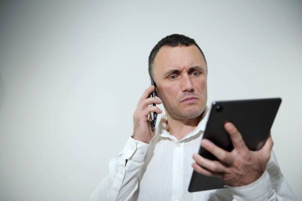 un homme avec un téléphone regardant avec indignation la tablette bougeait les sourcils sillonnés prix élevés changements de taux de change mauvais investissements problèmes au travail problèmes chemise blanche studio espace pour texte - back to front fashion men male photos et images de collection