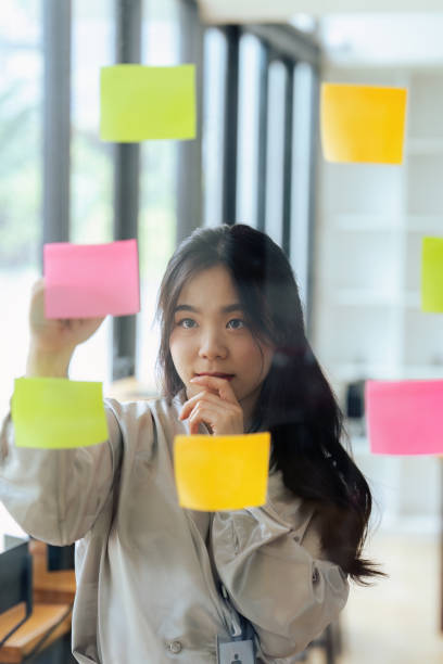 istock Through glass view of businesswoman analyzing process schedule, planning tasks on sticky papers 1635339391