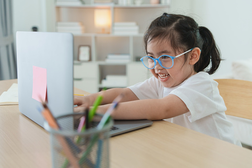 Asian baby girl wearing a white t-shirt and glasses use laptop and study online on wood table desk in living room at home. Education learning online from home concept.