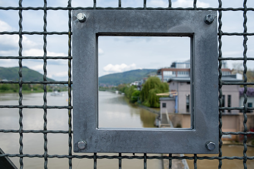 Steel wire mesh with view through square manufactured shaped hole at bridge over Neckar River, Heidelberg city, Germany. Blur building, nature, river water.