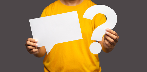 Close-up of hands young man holding a blank white speech bubble and a question mark symbol while standing on a gray background with copy space for text. Close-up photo