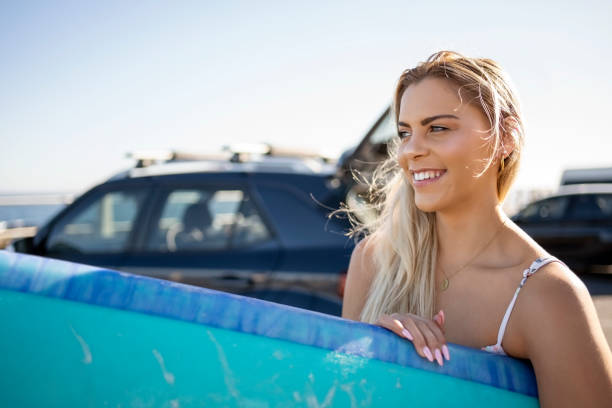 mulher jovem sorridente segurando sua prancha de surf em pé no estacionamento da praia - beach parking lot car equipment - fotografias e filmes do acervo