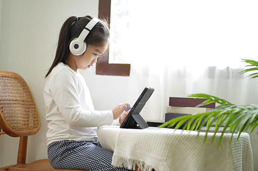 Little Asian girl sitting on the chair nearby window in the moring light, Choosing music playlist on digital tablet and listen music on wireless headphones