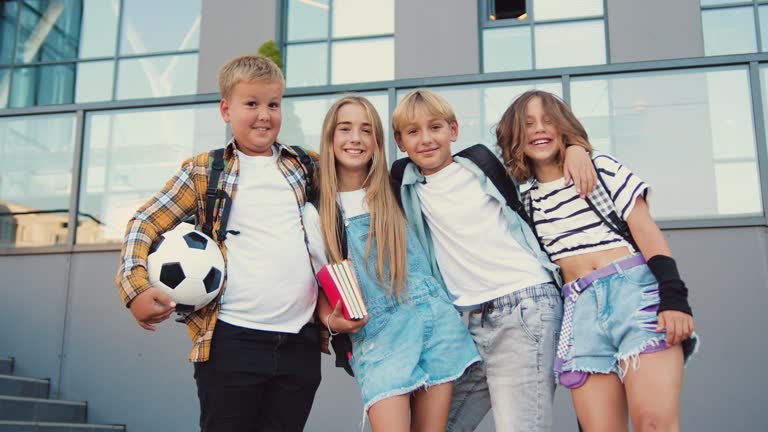 Portrait happy smiling group of high school kids hugging looking at camera. Children two girls and two boys pose standing on the steps of a modern school building in the background.