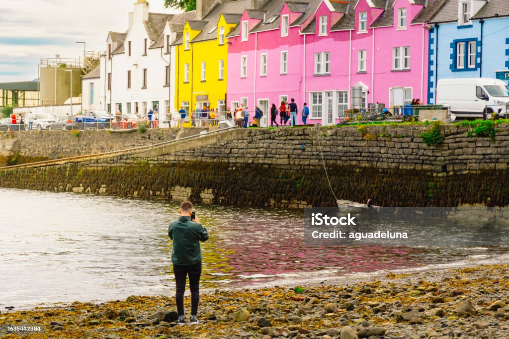 Unrecognisable young man taking a photo with a smartphone of the colourful houses at Portree, Scotland. Unrecognisable young man taking a photo with a smartphone of the colourful houses at Portree, Scotland. Horizontal shot. Architecture Stock Photo