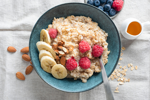 Oatmeal porridge in blue bowl topped with raspberry, banana and almond nuts. Table top view. Healthy vegan breakfast meal