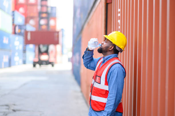 istock African American man logistic staff workers wearing reflective vests and white helmets in shipping container yard, Drinking water when resting after working. Cargo Ship Import Export Factory Logistic. 1635511226