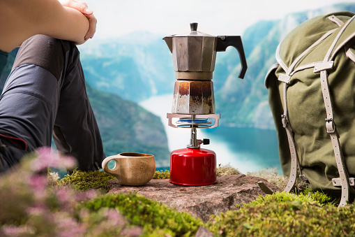 Cooking, making coffee or tea on a portable camping gas burner with a red gas cylinder with a Scandinavian nature background. Summer hiking, ecotourism, survival.