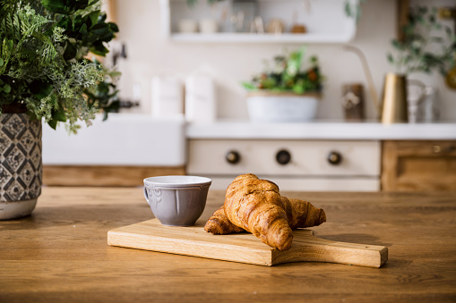 closeup of french croissants and cup with coffee on wooden cutting board on blurred kitchen background. fresh pastry with tea and vase with flowers on dining table at home. delicious breakfast concept