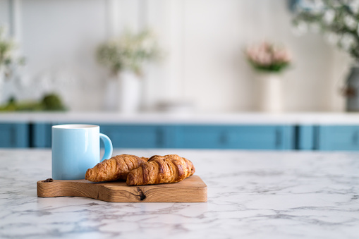 Wooden kitchen board with croissants and blue coffee mug on stone marble table top. Blurred abstract background from interior. Concepts of tasty breakfast, copy space.