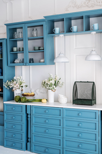 Vertical shot of blue wooden furniture in stylish kitchen. A lot of storage drawers for utensils. Fresh flowers on countertop. Vintage decor elements against white wall