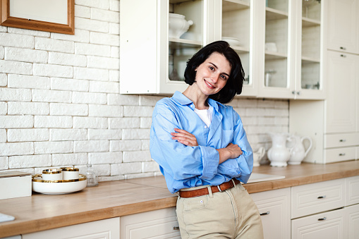 Portrait of young successful self employed woman in blue shirt standing with crossed arms at home kitchen, looking at camera and smile. Confident female freelancer in casual clothes in apartment