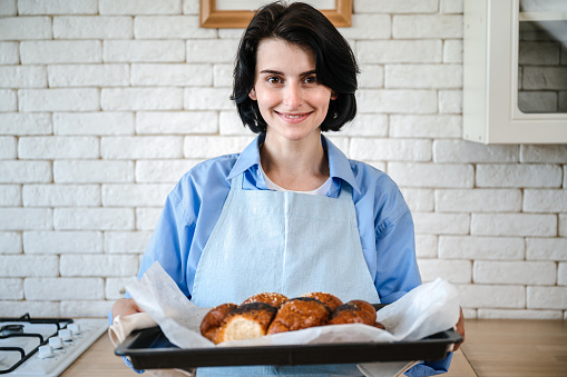 Portrait of happy woman in apron holding tray with fresh homemade bakery at modern kitchen. Smelling appetizing chrunchy gluten free bun from flour and seeds.